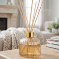 champagne glass reed diffuser with a gold collar and natural wood reeds, displayed on a wooden table in a cozy living room setting.