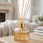 champagne glass reed diffuser with a gold collar and natural wood reeds, displayed on a wooden table in a cozy living room setting.