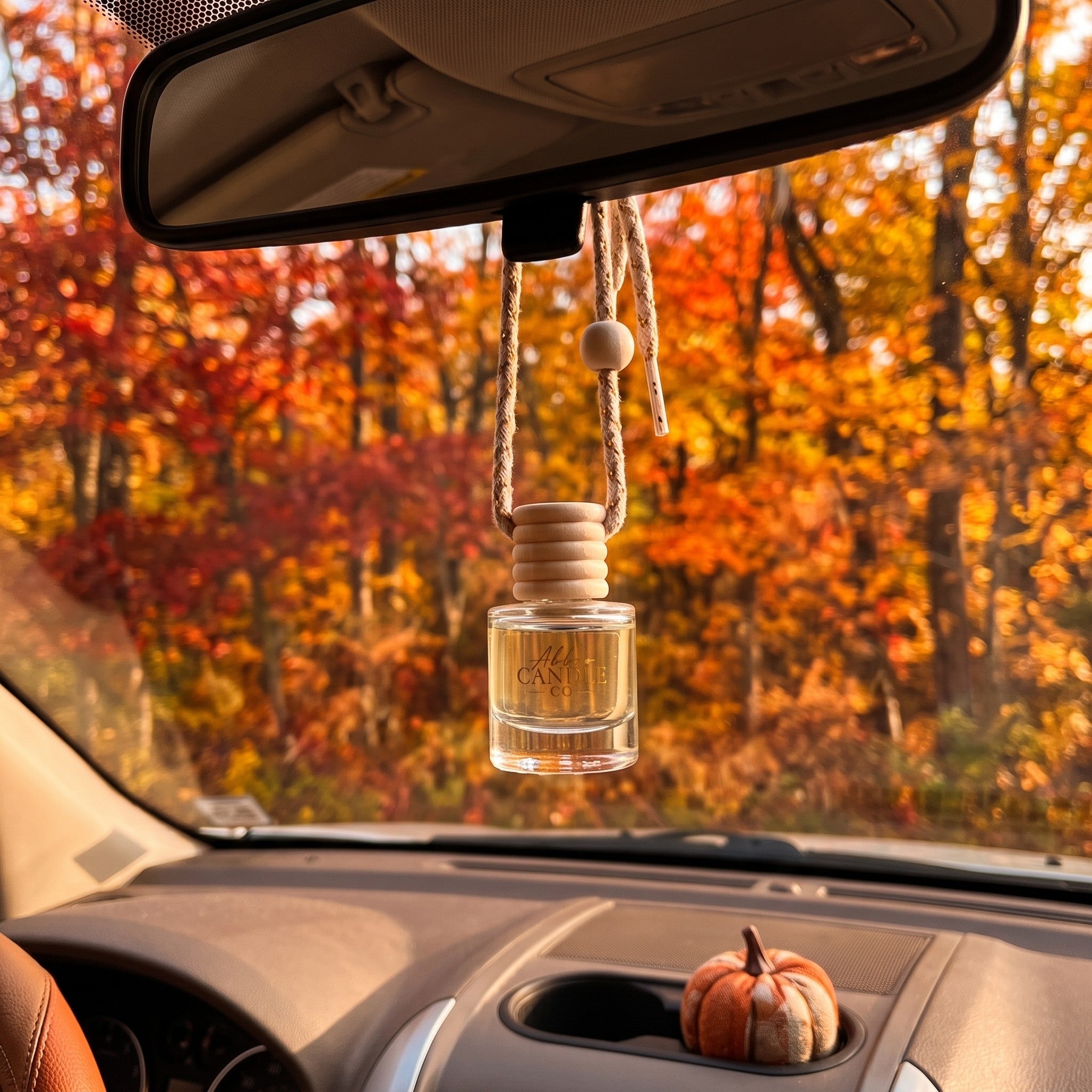A clear glass car air freshener bottle with a light wood diffuser top hangs from a rope under a rearview mirror inside a car. The background shows a blurred autumn forest with bright orange and red foliage, and a small orange pumpkin sits in the car's cup holder.
