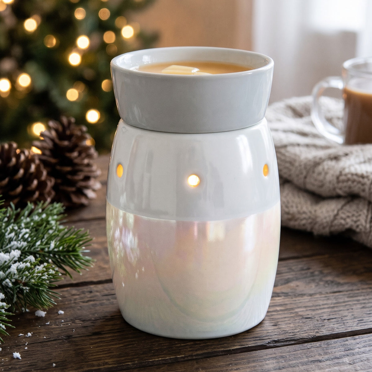 A ceramic wax warmer with a light gray top section and a pearlescent white lower body, lit with warm internal lights. It sits on a rustic wooden table with a blurred Christmas tree, pinecones, evergreen branches, and a mug of coffee in the background.