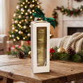 A white rectangular gift box with a clear window displaying stacked gold tins, tied with a dark green ribbon, set on a wooden table in a cozy holiday living room with a Christmas tree and fireplace in the background.