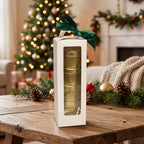 A white rectangular gift box with a clear window displaying stacked gold tins, tied with a dark green ribbon, set on a wooden table in a cozy holiday living room with a Christmas tree and fireplace in the background.