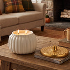 A white ribbed pumpkin-shaped candle with two lit wicks sits on a rustic wooden table in a cozy living room setting, alongside a gold lid on a coaster.