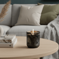 A lit dark amber glass candle sits on a light wood coffee table in a cozy living room setting with a gray sofa, decorative pillows, a small vase, and books.