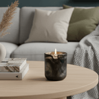 A lit dark amber glass candle sits on a light wood coffee table in a cozy living room setting with a gray sofa, decorative pillows, a small vase, and books.