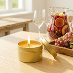 Yellow candle in a tin on a table with a pitcher of fruit punch and grapes in the background.