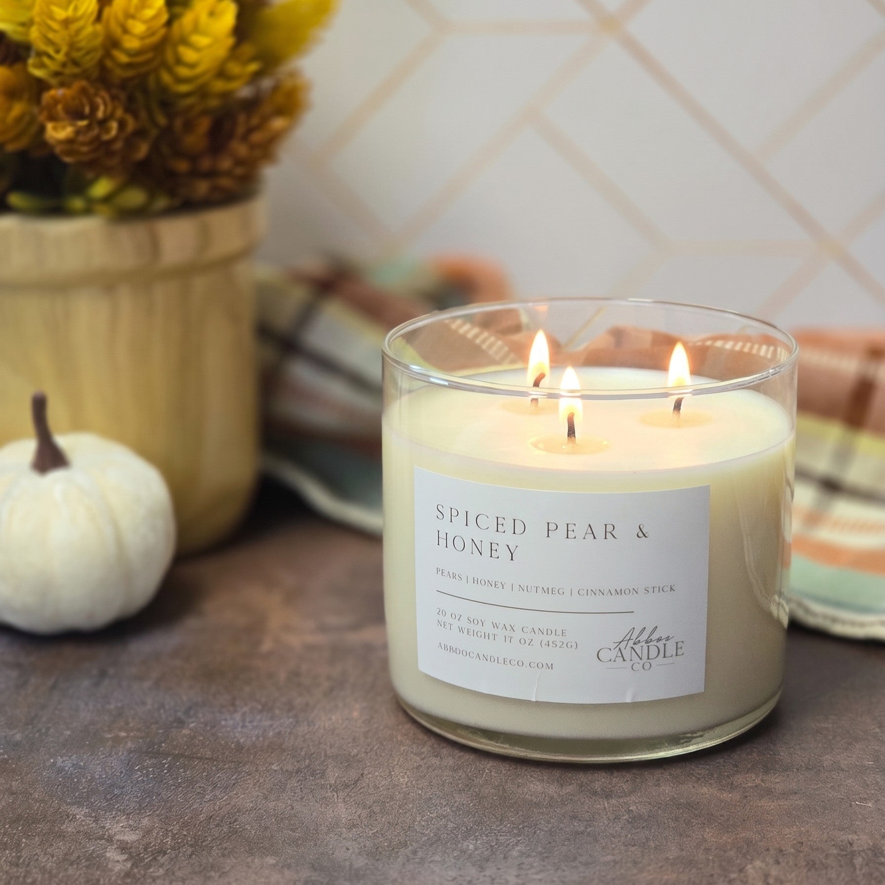 A lit 3-wick candle in a clear glass jar sits on a rustic tabletop. The label reads 'Spiced Pear & Honey' and shows the brand Abboo Candle Co.