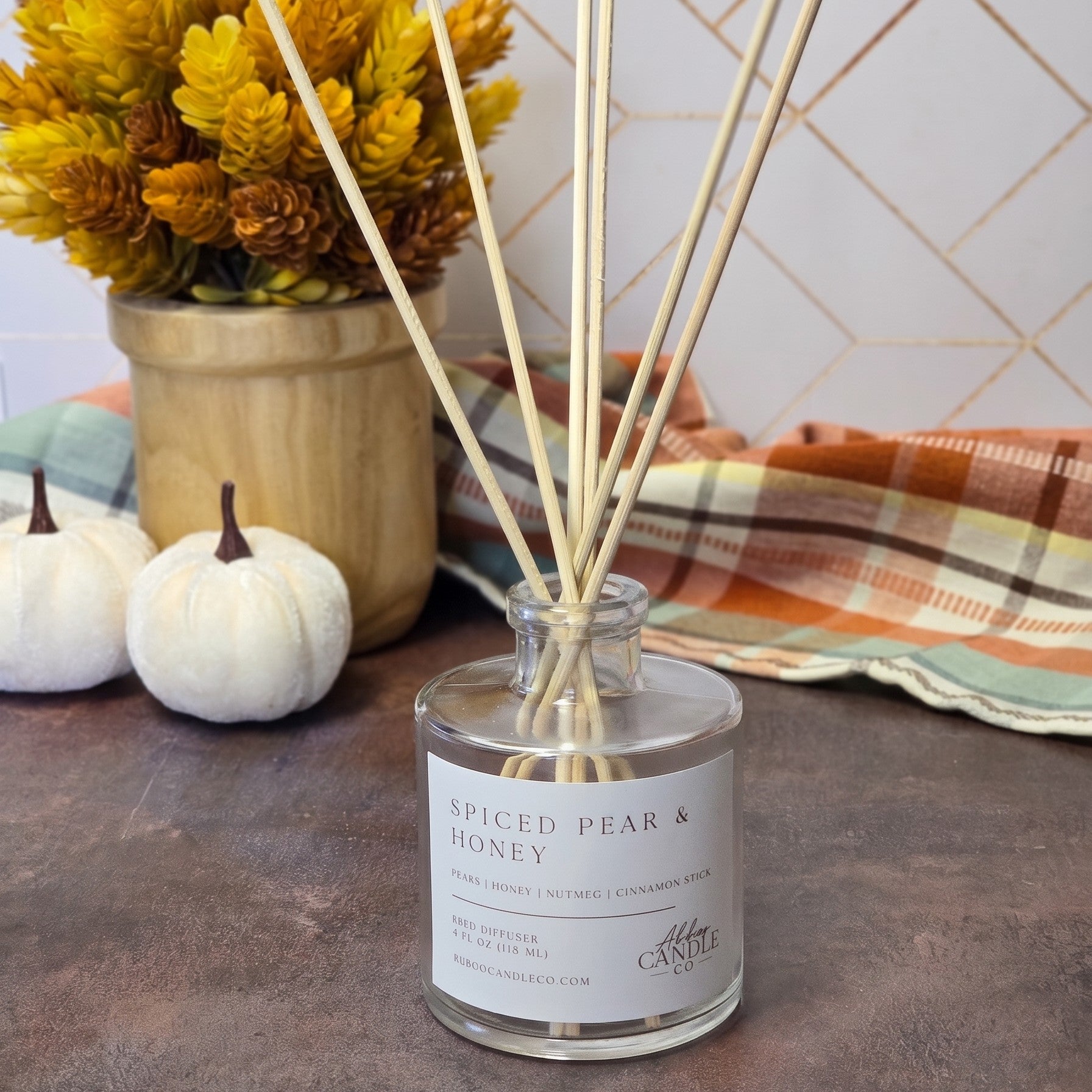 A clear glass reed diffuser bottle labeled 'Spiced Pear & Honey' with natural reed sticks, styled on a tabletop with autumn decor including pumpkins, a plaid cloth, and dried yellow flowers.
