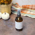 An amber glass spray bottle of Spiced Pear Honey with a white label and clear spray cap, set on a rustic brown surface with a plaid blanket and small pumpkins in the background.