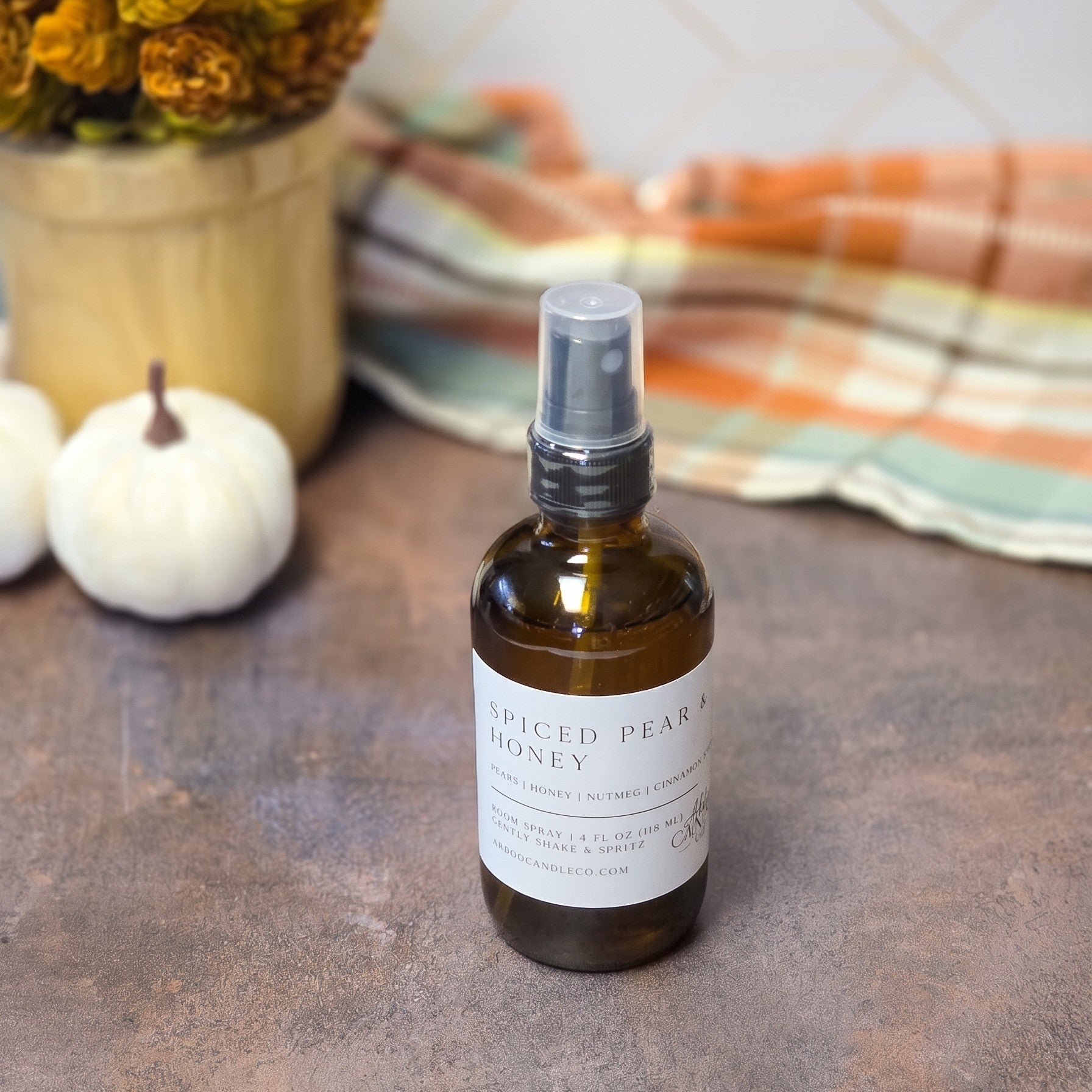 An amber glass spray bottle of Spiced Pear Honey with a white label and clear spray cap, set on a rustic brown surface with a plaid blanket and small pumpkins in the background.
