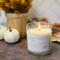 A lit scented candle in a clear glass jar sits on a rustic surface with a white label reading 'Spiced Pear & Honey' and the brand name 'Aromatic Candle Co.' Background includes pumpkins, a vase of dried flowers, and a tiled wall, creating a warm home setting.