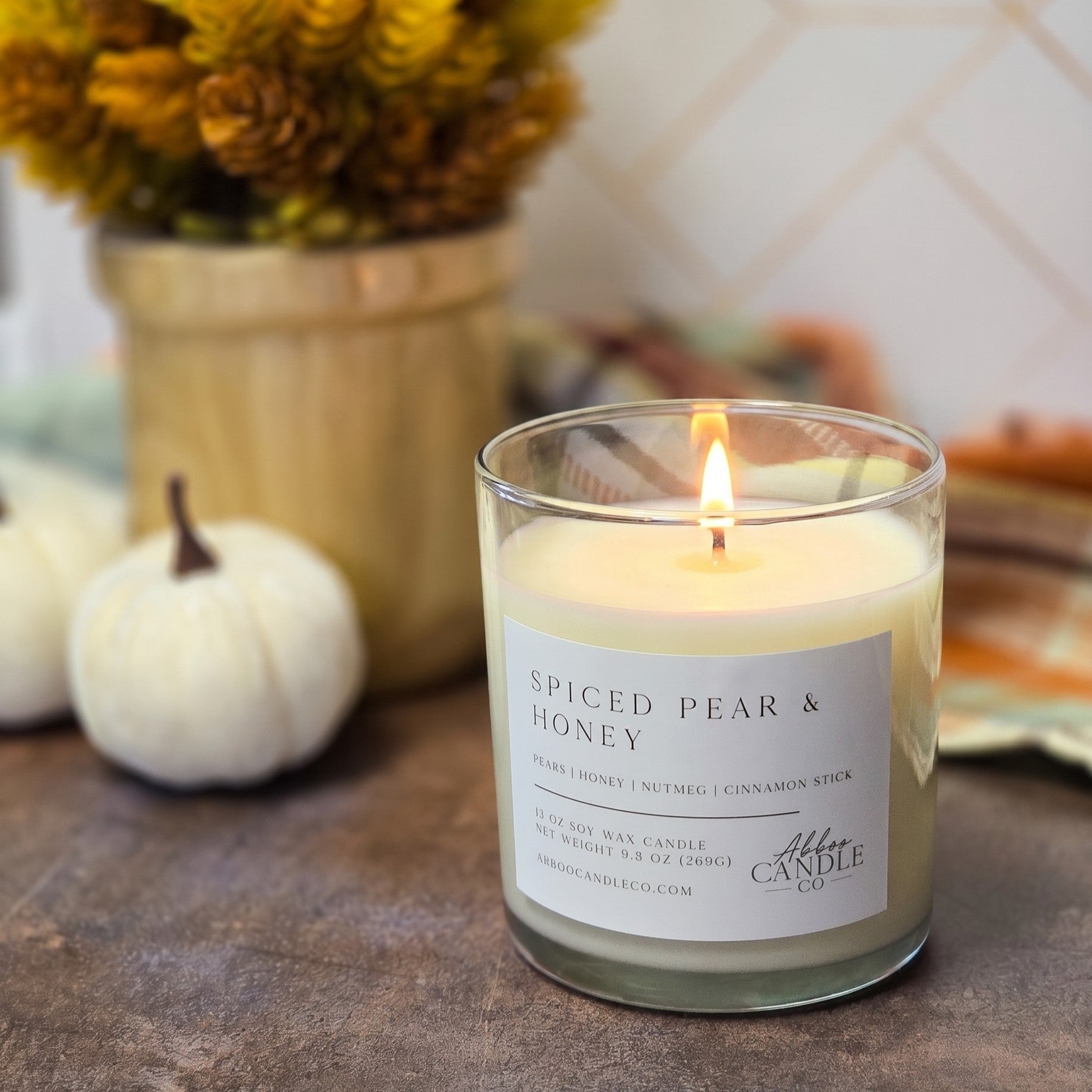 A lit scented candle in a clear glass jar sits on a rustic surface with a white label reading 'Spiced Pear & Honey' and the brand name 'Aromatic Candle Co.' Background includes pumpkins, a vase of dried flowers, and a tiled wall, creating a warm home setting.