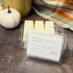 A clear plastic clamshell package of wax melts labeled 'Spiced Pear & Honey' by Abbot Candle Co, placed on a dark tabletop with a small white pumpkin and warm-toned background decor.