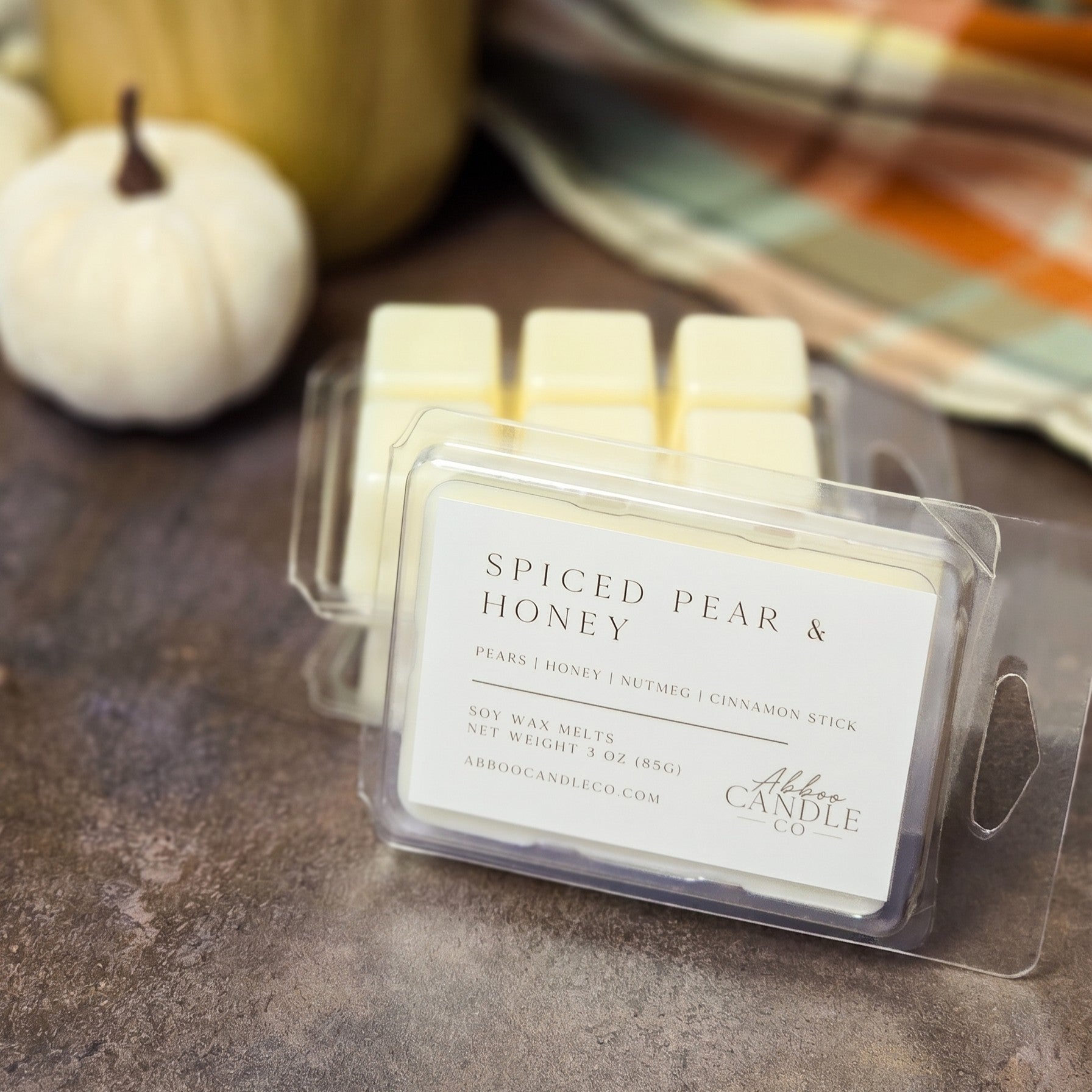 A clear plastic clamshell package of wax melts labeled 'Spiced Pear & Honey' by Abbot Candle Co, placed on a dark tabletop with a small white pumpkin and warm-toned background decor.