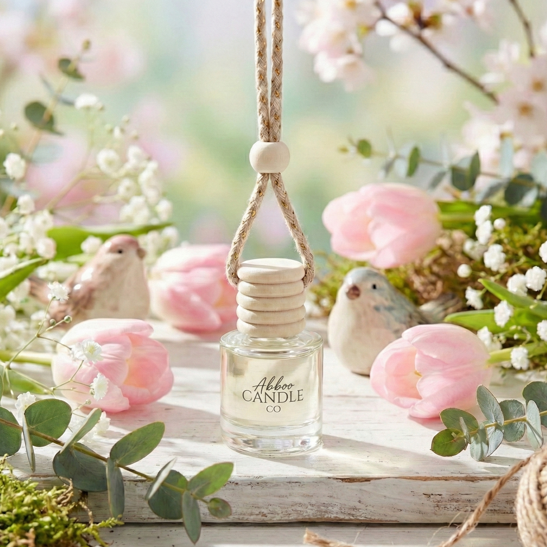 A bottle of Abboo Candle Co. hanging car diffuser on a wooden surface with pink flowers and greenery.
