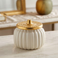 A ribbed white ceramic jar with a textured gold lid, set on a light marble surface with a softly blurred background.