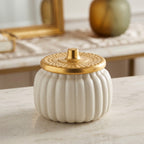 A ribbed white ceramic jar with a textured gold lid, set on a light marble surface with a softly blurred background.
