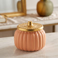 A ribbed ceramic pumpkin-shaped container in a warm peach-pink color with a metallic gold lid, set on a light marble surface.