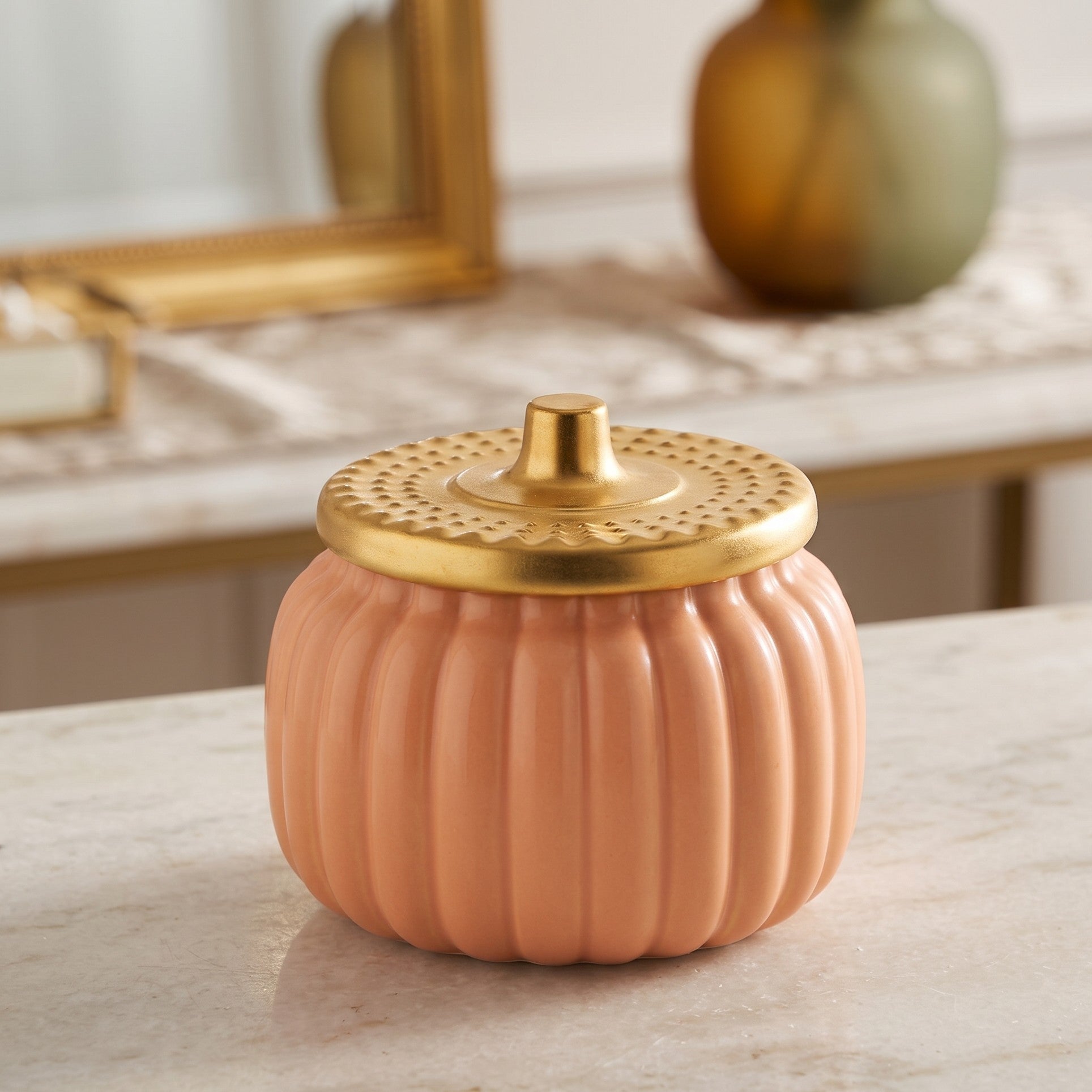 A ribbed ceramic pumpkin-shaped container in a warm peach-pink color with a metallic gold lid, set on a light marble surface.