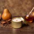A small gold metal candle tin with a lit candle sits on a wooden table, with its lid leaning beside it. The scene is styled with a pear, cinnamon sticks, star anise, and a glass jar of honey in the background, creating a warm, cozy atmosphere.