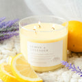 A clear glass soy candle with three wicks, labeled 'Lemon and Lavender', surrounded by lemon slices and lavender flowers.